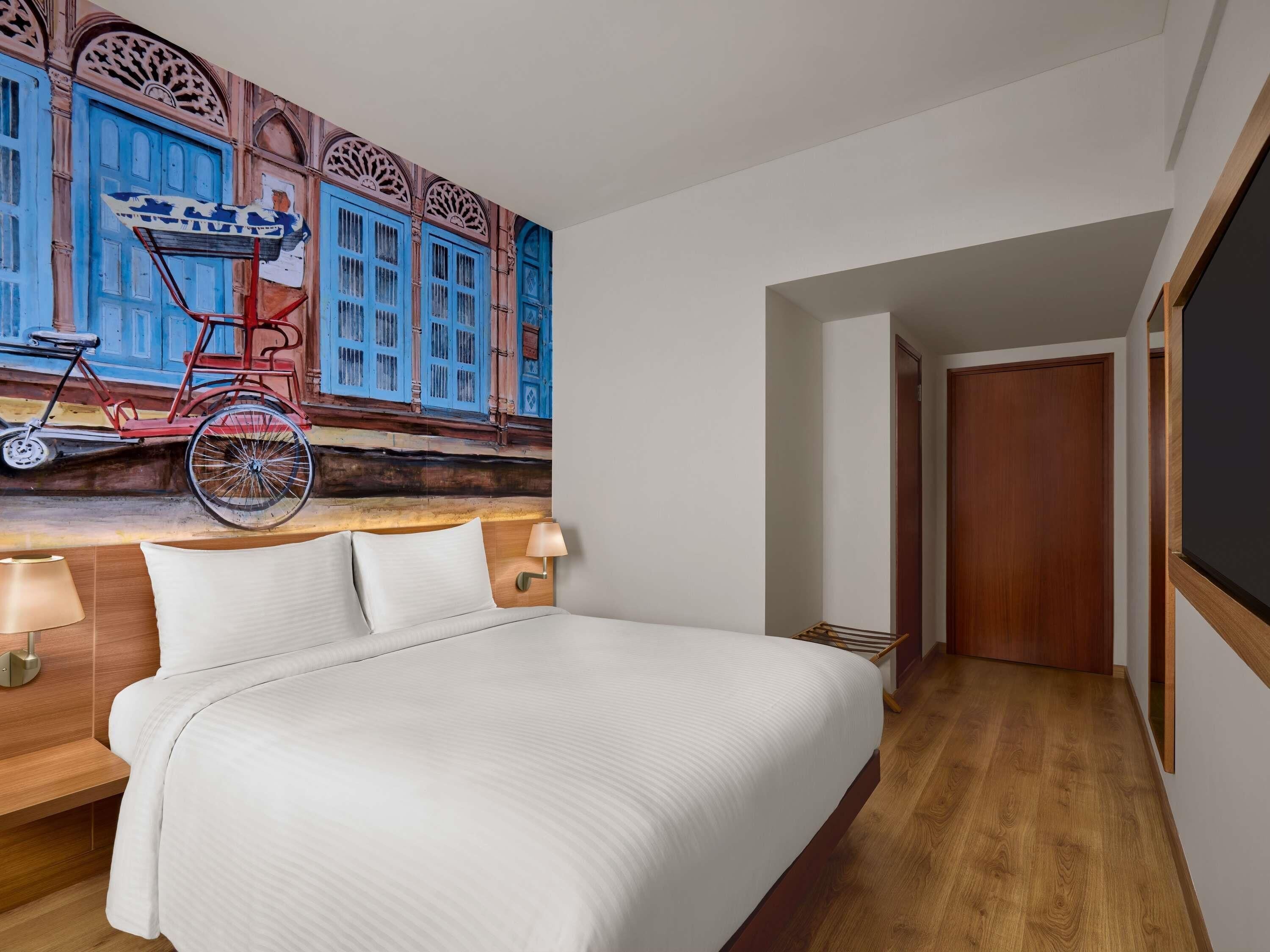 A hotel room with a bed, a wall-mounted lamp, a wooden headboard, and a mural of a bicycle.
