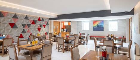 A modern dining area with multiple tables and chairs, a kitchen in the background, and a colorful wall mural.
