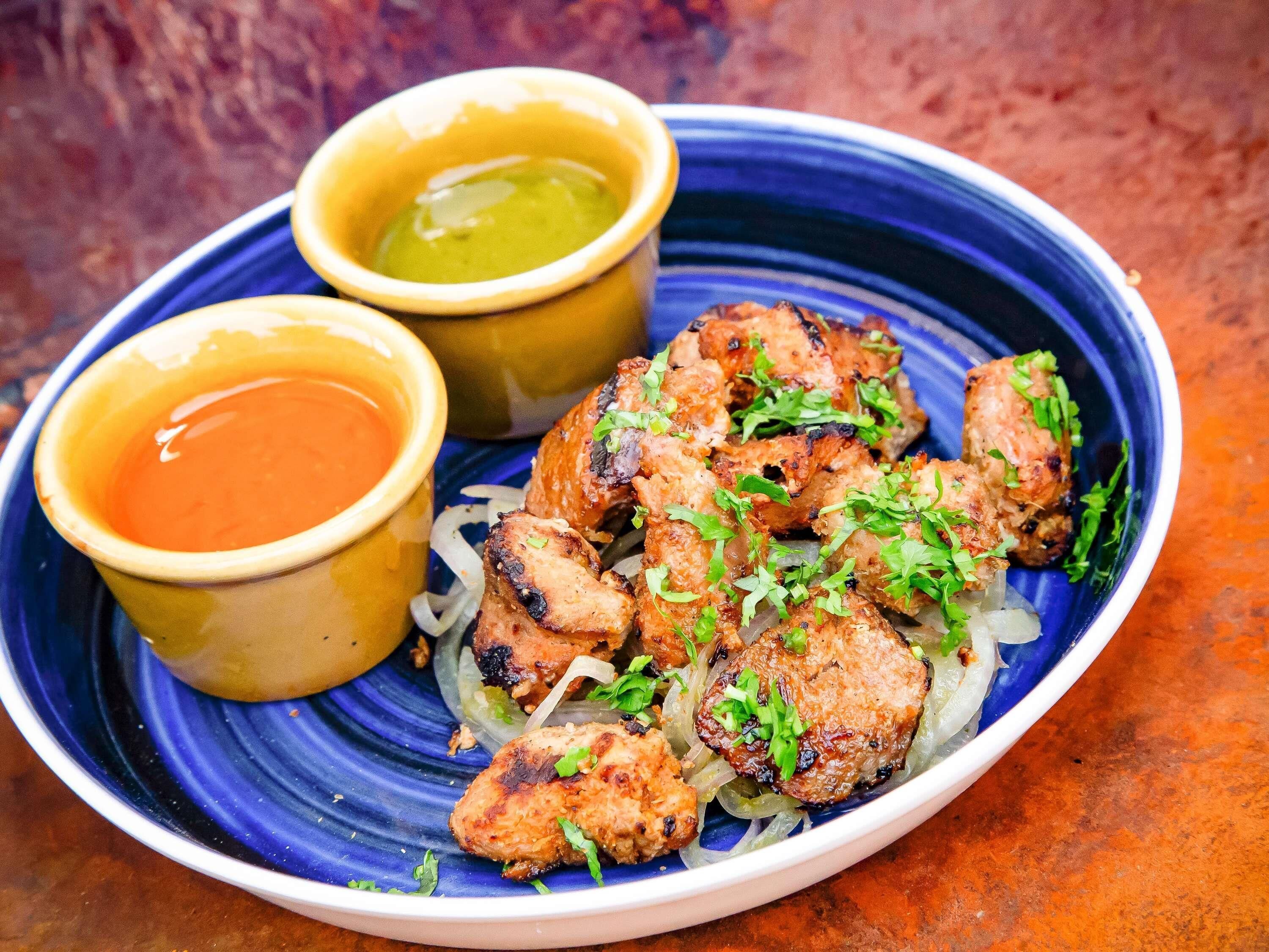 A plate of grilled meat with onions and garnished with herbs, accompanied by two small bowls of sauces.