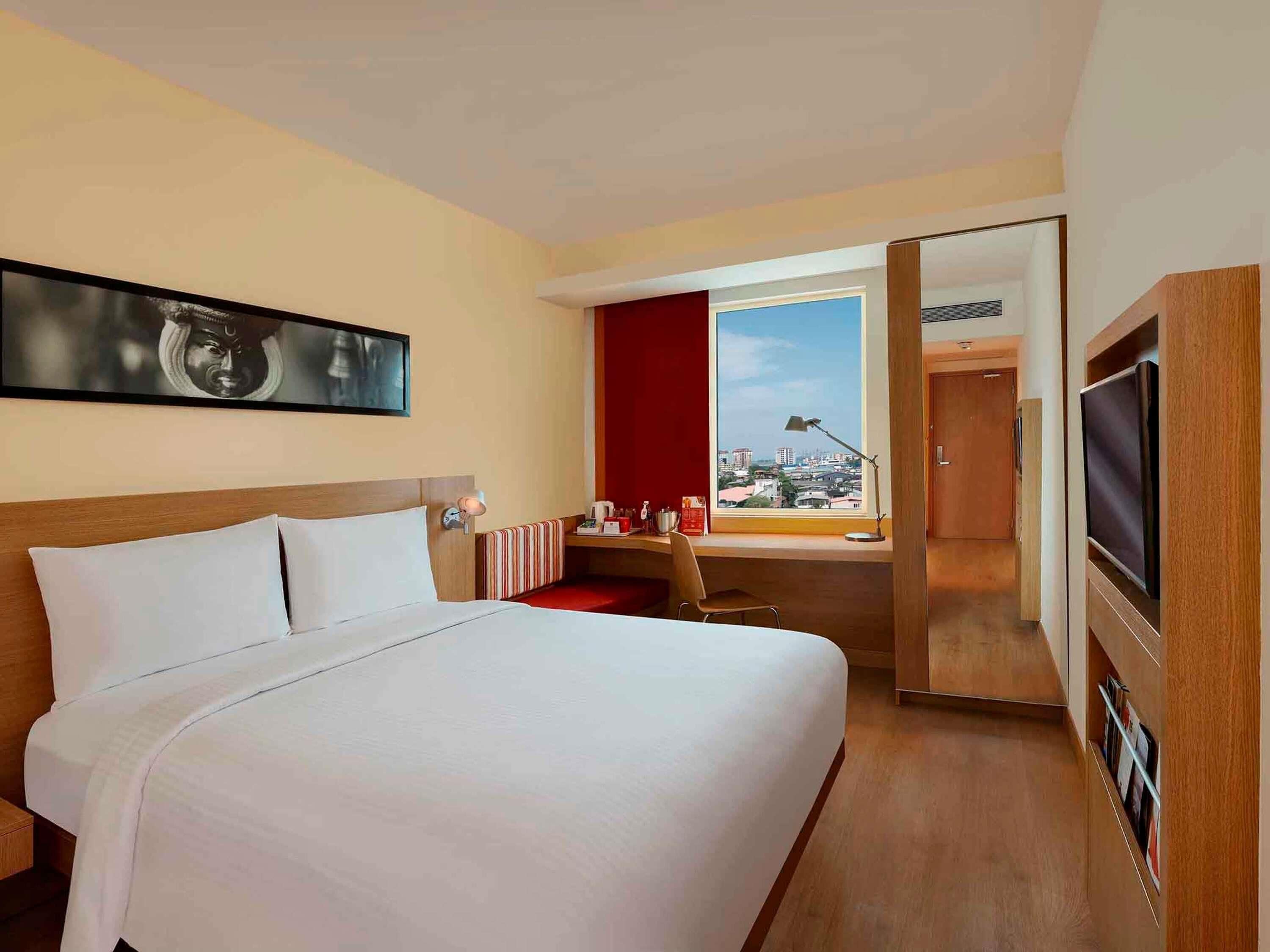 Une chambre d’hôtel avec un grand lit, un bureau avec une chaise, une télévision et une fenêtre donnant sur la ville.