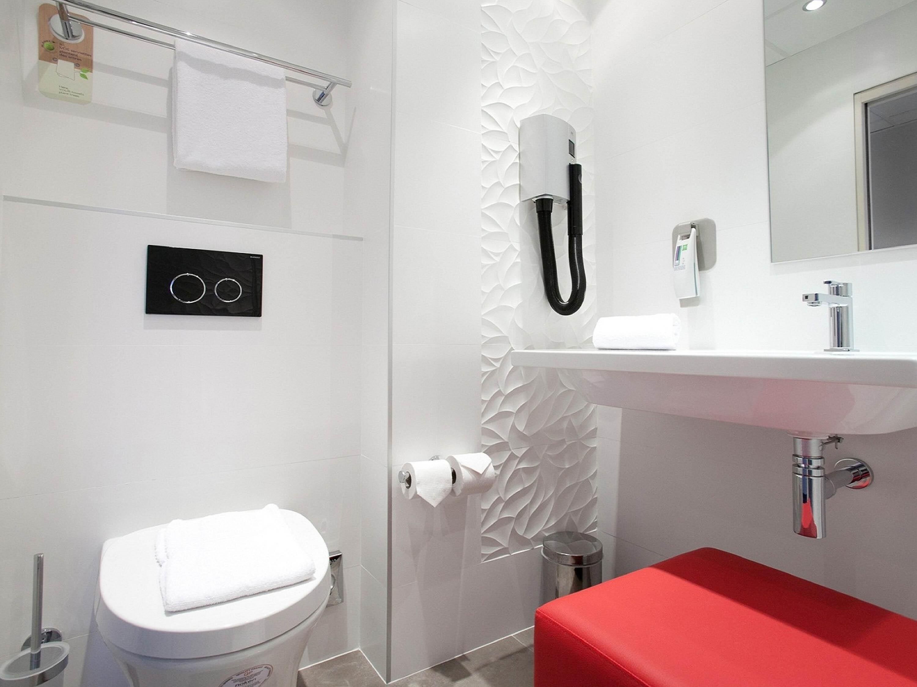 A modern bathroom with a white toilet, a red bench, a white sink, a mirror, and a towel rack.