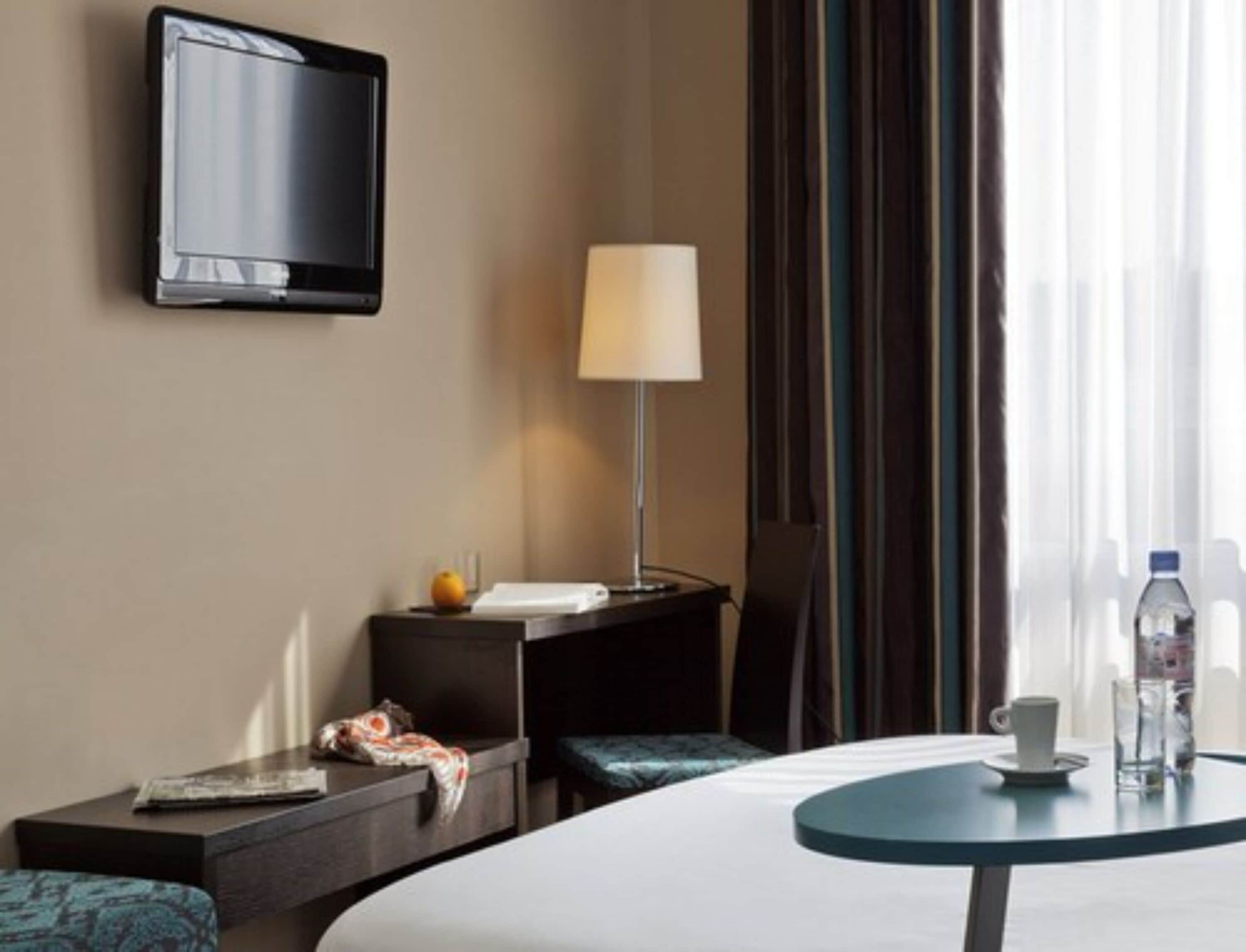 A hotel room with a bed, a bedside table with a lamp, a TV, a small table with a cup and water bottle, and a window with curtains.