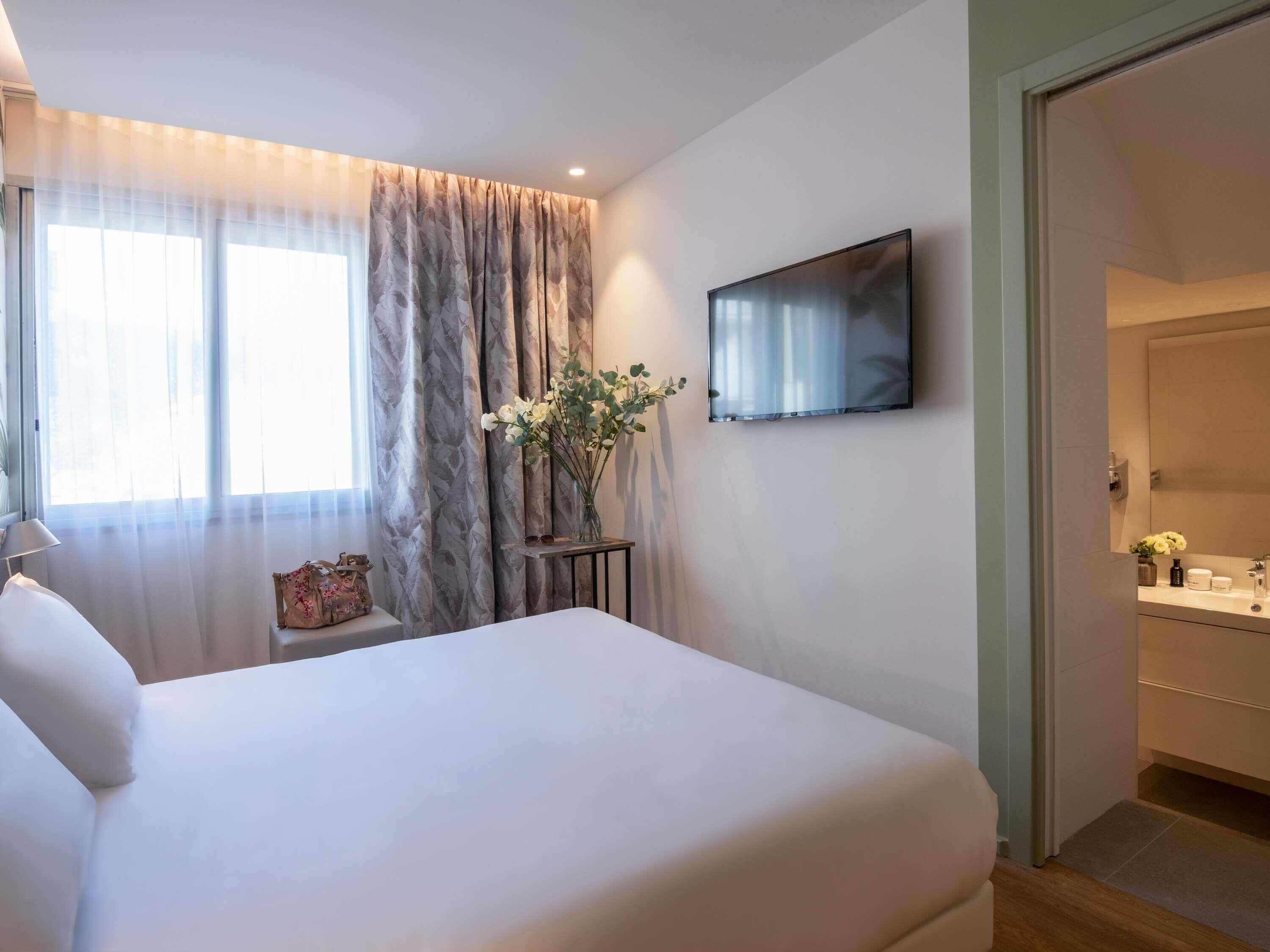 A bedroom with a bed, a TV mounted on the wall, a bedside table with a vase of flowers, and a window with sheer curtains.