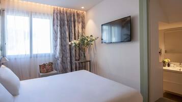 A bedroom with a bed, a TV mounted on the wall, a bedside table with a vase of flowers, and a window with sheer curtains.