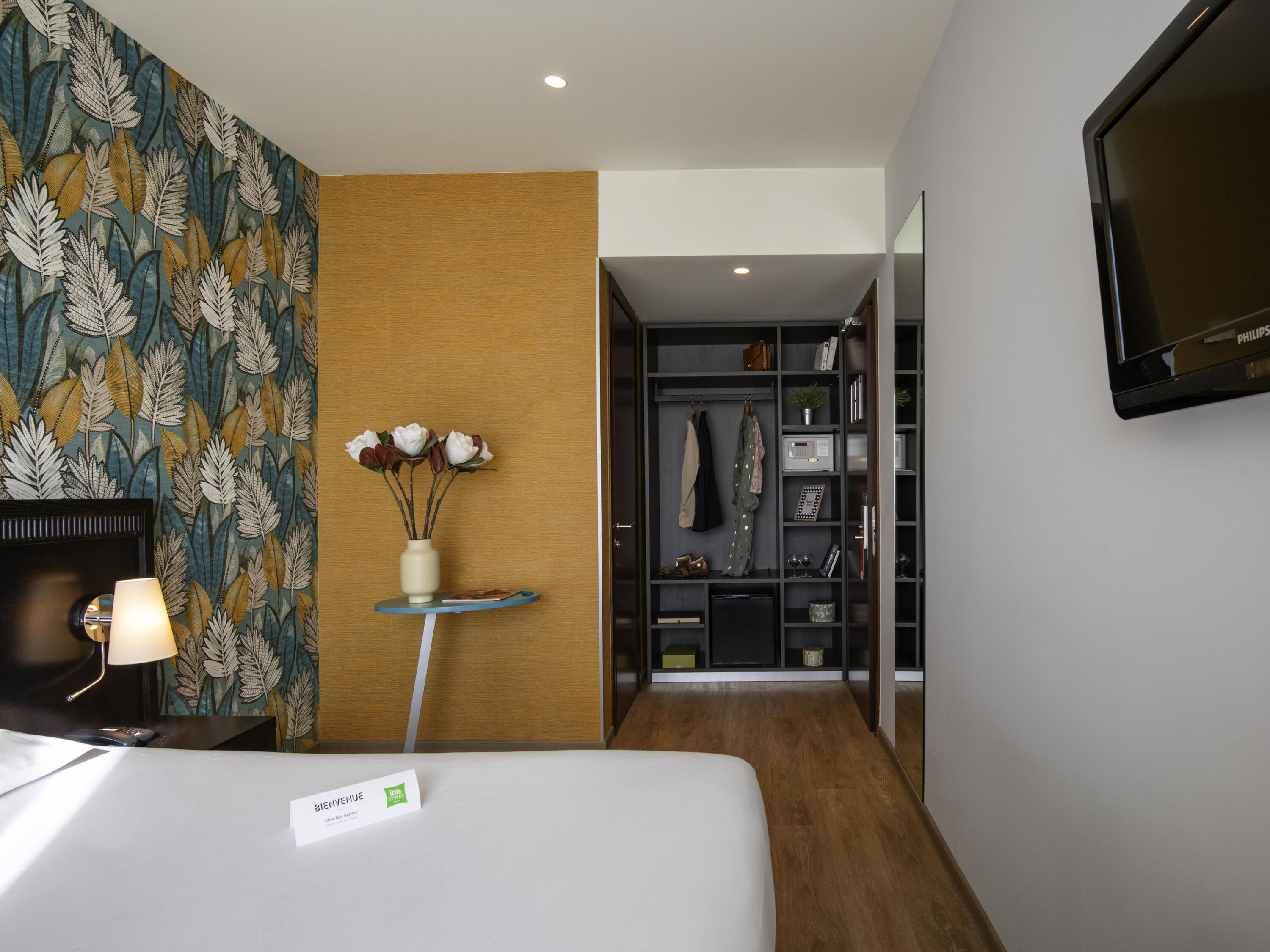A hotel room with a bed, a desk, a lamp, a vase with flowers, a wardrobe, and a television.