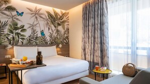A hotel room with a bed, a bedside table with breakfast items, a sofa, a small round table with a book and mug, a large window with sheer curtains, and a wall with a tropical leaf mural. - ibis Styles Beaulieu Sur Mer (Beaulieu-sur-Mer)