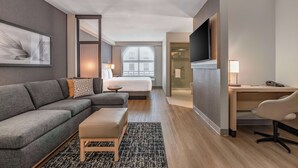 A modern hotel room with a grey sofa, a small wooden table, a desk with a chair, a TV mounted on the wall, and a large window with a view of the city. - Hyatt Place Riverside Downtown (Riverside)