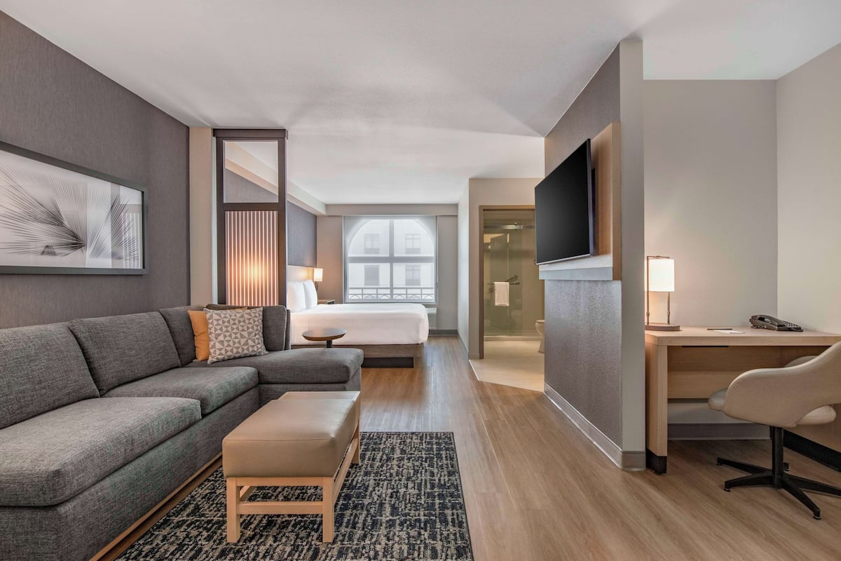 A modern hotel room with a grey sofa, a small wooden table, a desk with a chair, a TV mounted on the wall, and a large window with a view of the city.