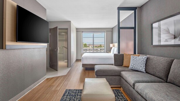A modern hotel room with a large bed, a sofa, a flat-screen TV, and a balcony with a city view. - Hyatt Place Riverside Downtown (Riverside)