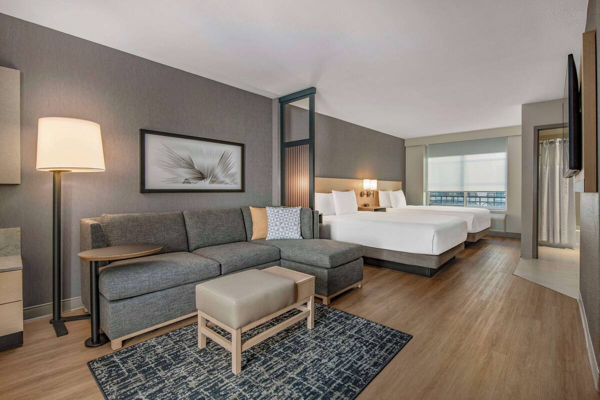 A modern hotel room with a grey sofa, a small table, a bed with white linens, a large window, and a flat-screen TV mounted on the wall.