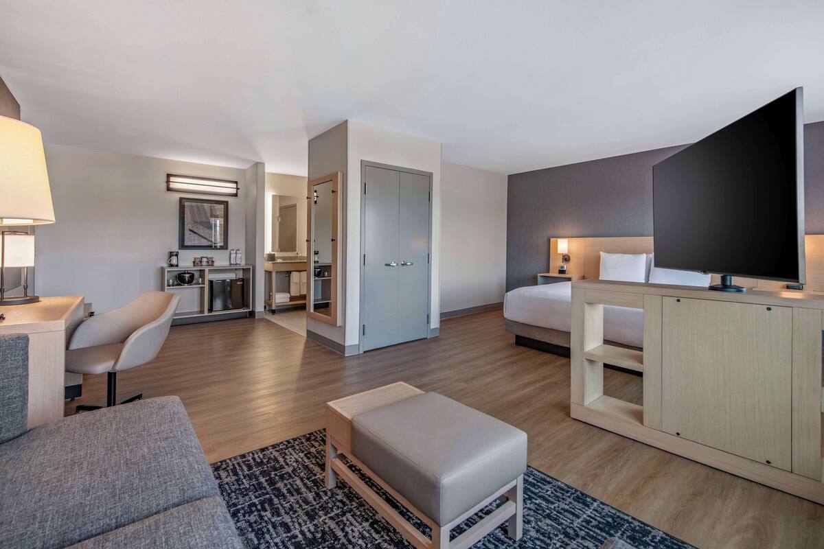 A modern hotel room with a sofa, a small table, a TV, a bed, a desk, and a kitchenette.