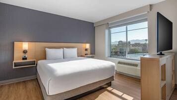 A modern hotel room with a large bed, bedside tables with lamps, a TV, and a large window with a city view.