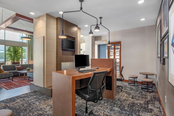 A modern office space with a wooden desk, black office chair, and a computer. There is a television mounted on the wall, a sofa, and a small seating area with stools.