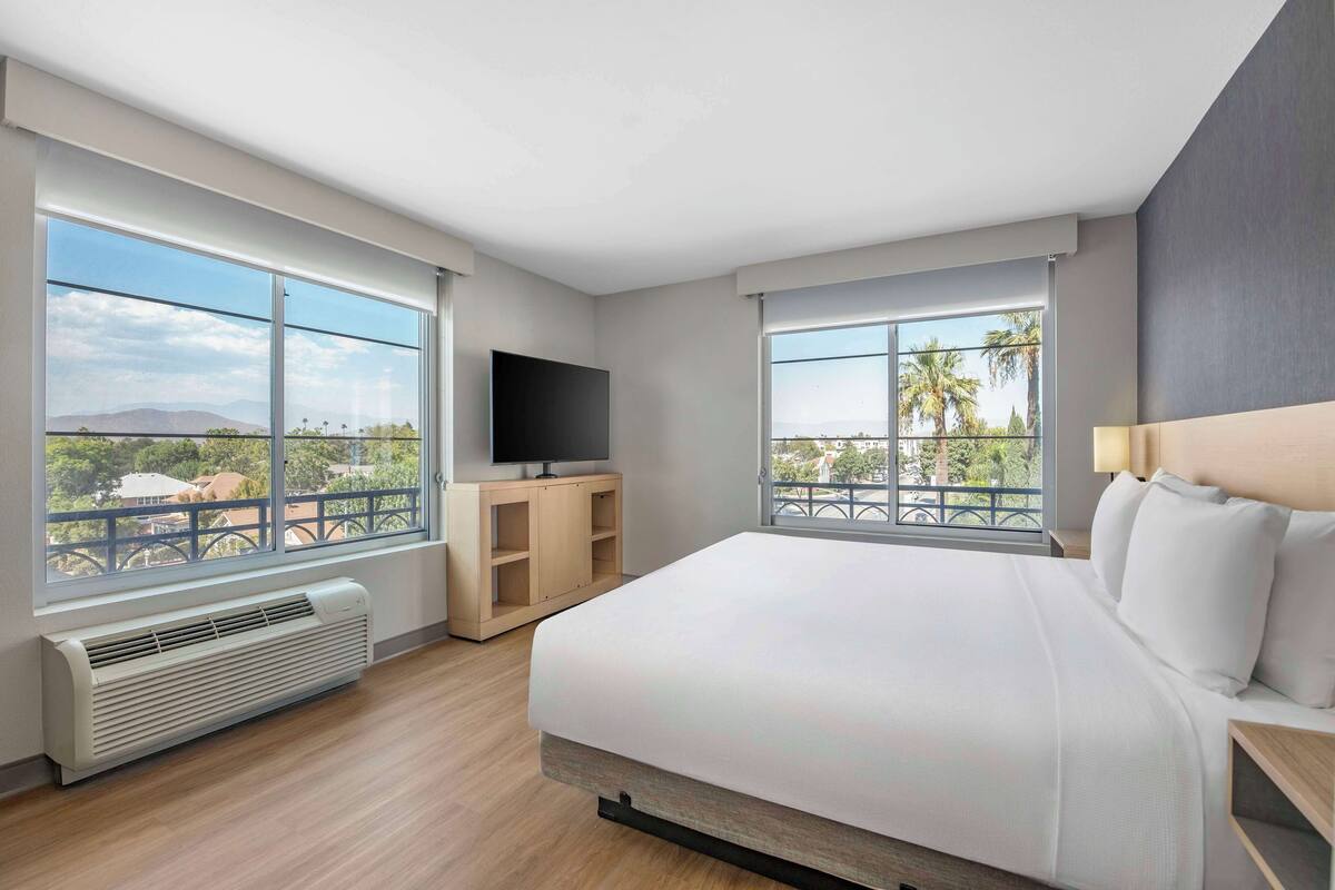 A modern hotel room with a large bed, a flat-screen TV, a heater, and a balcony with a view of palm trees and mountains.