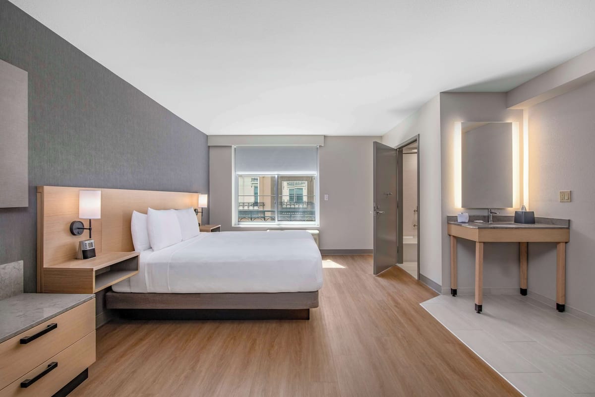A modern hotel room with a large bed, bedside table, lamp, and a bathroom with a vanity and mirror.