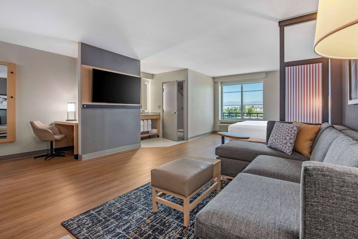 A modern hotel room with a sofa, ottoman, desk, and TV.