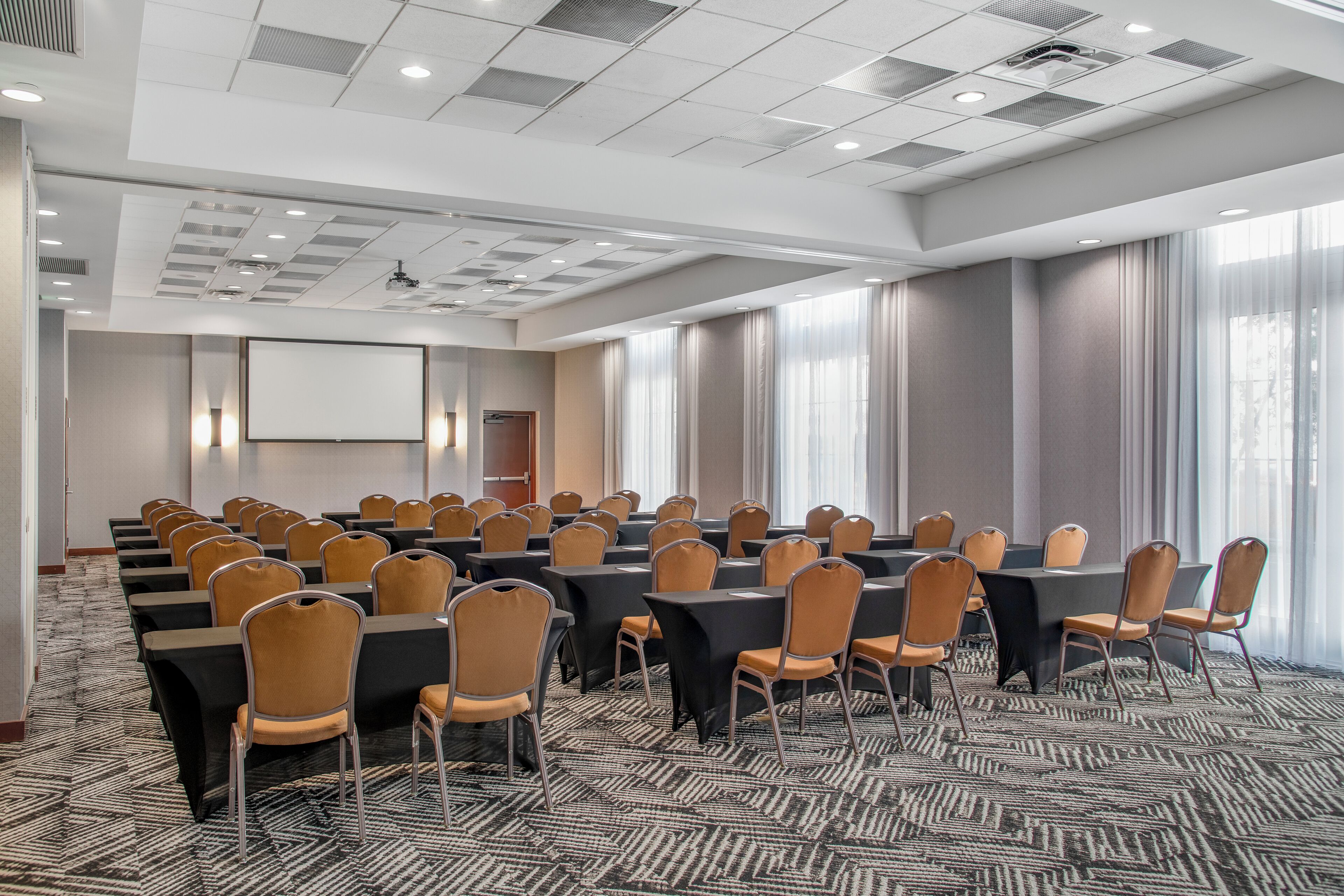 A conference room with multiple tables and chairs, a projector screen, and large windows with curtains.
