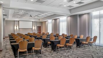 A conference room with multiple tables and chairs, a projector screen, and large windows with curtains.
