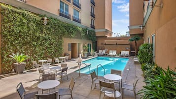 A poolside area with lounge chairs and tables, surrounded by a building with a climbing vine-covered wall and a clear blue pool.