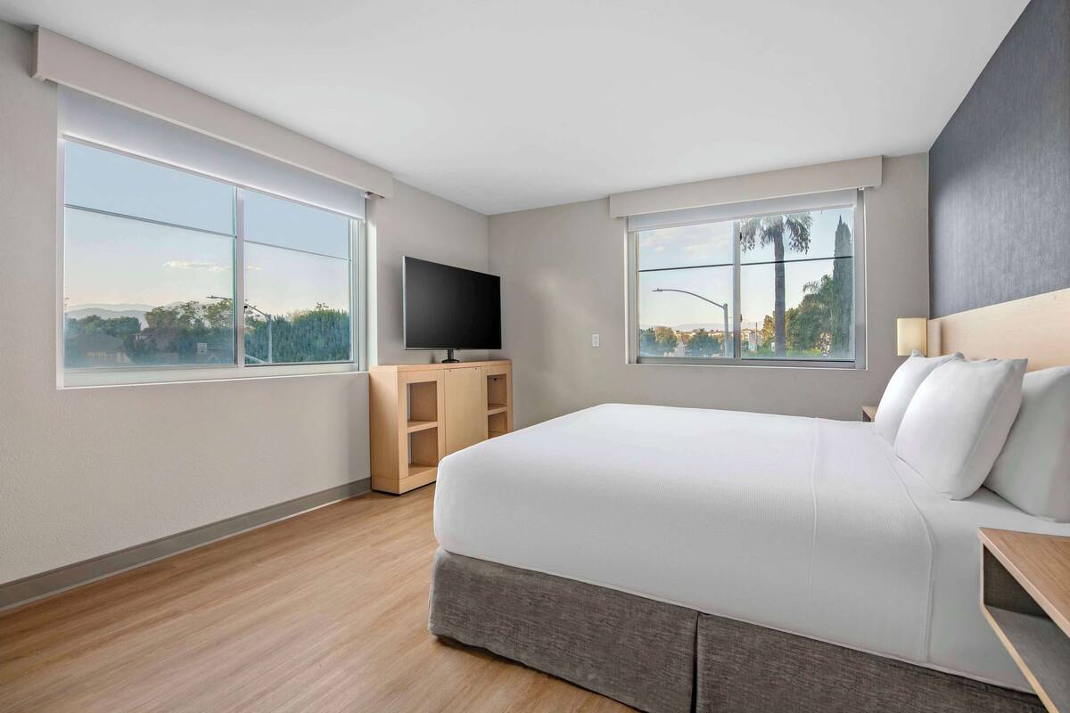 A modern hotel room with a large bed, a flat-screen TV, a wooden cabinet, and large windows offering a view of the outdoors.