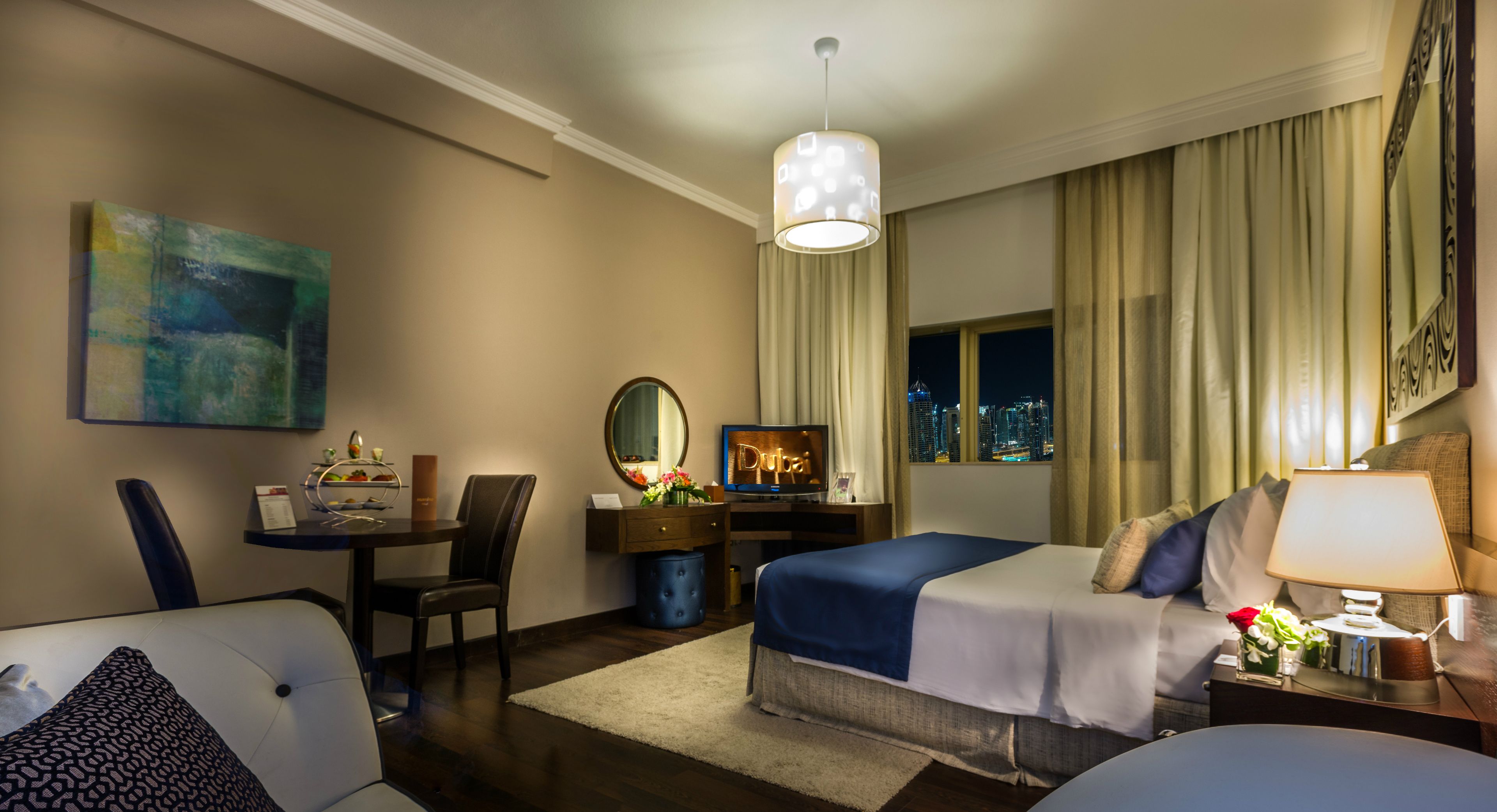 Une chambre d’hôtel avec un lit, un bureau avec une chaise, une télévision, une lampe de bureau et une vue sur la ville la nuit.