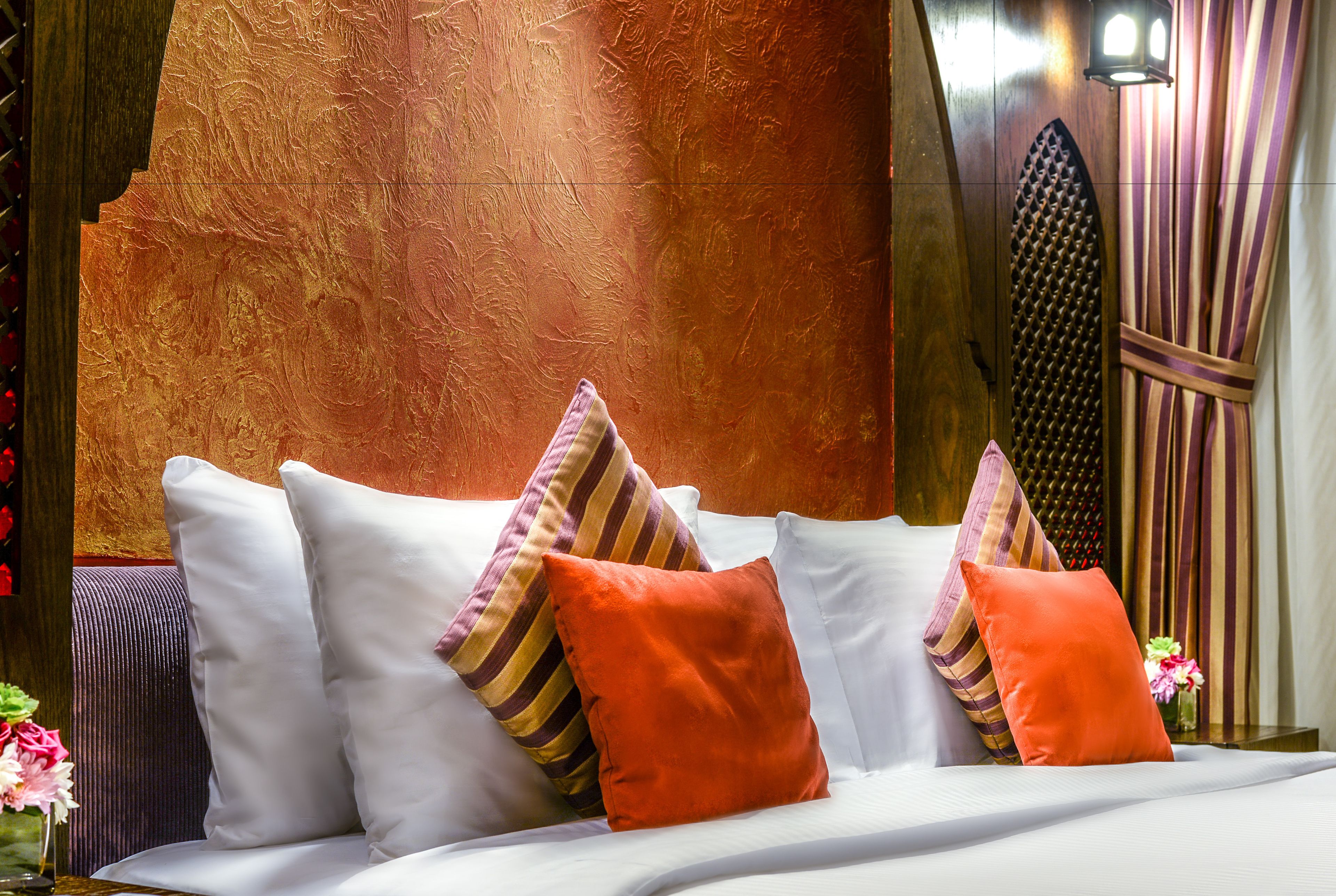 A neatly made bed with white linens and multiple pillows, including striped and solid orange ones, in a room with a textured wall and a decorative wall panel.