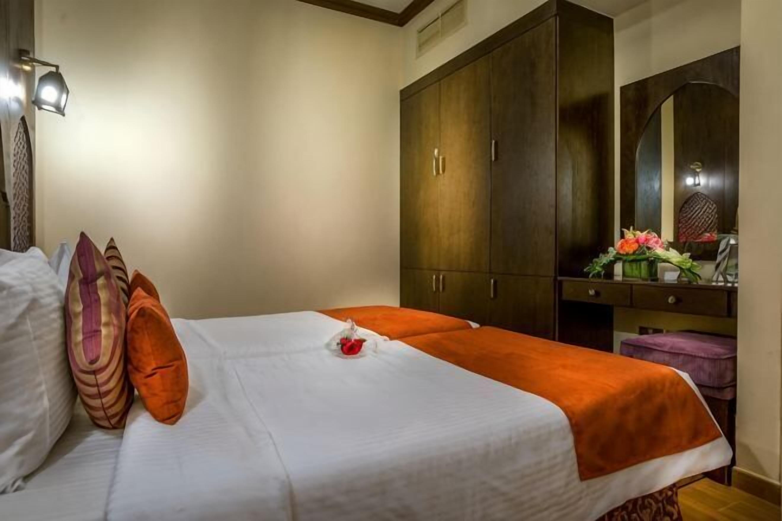 Une chambre d’hôtel comprenant un lit, une armoire, une coiffeuse avec un miroir et une petite table sur laquelle est posé un vase de fleurs.