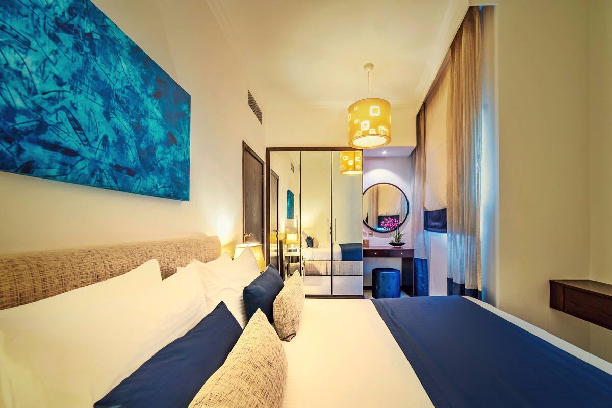 Ein modernes Hotelzimmer mit einem großen Bett, einem Spiegel, einem Schreibtisch und einem blauen, abstrakten Gemälde an der Wand.