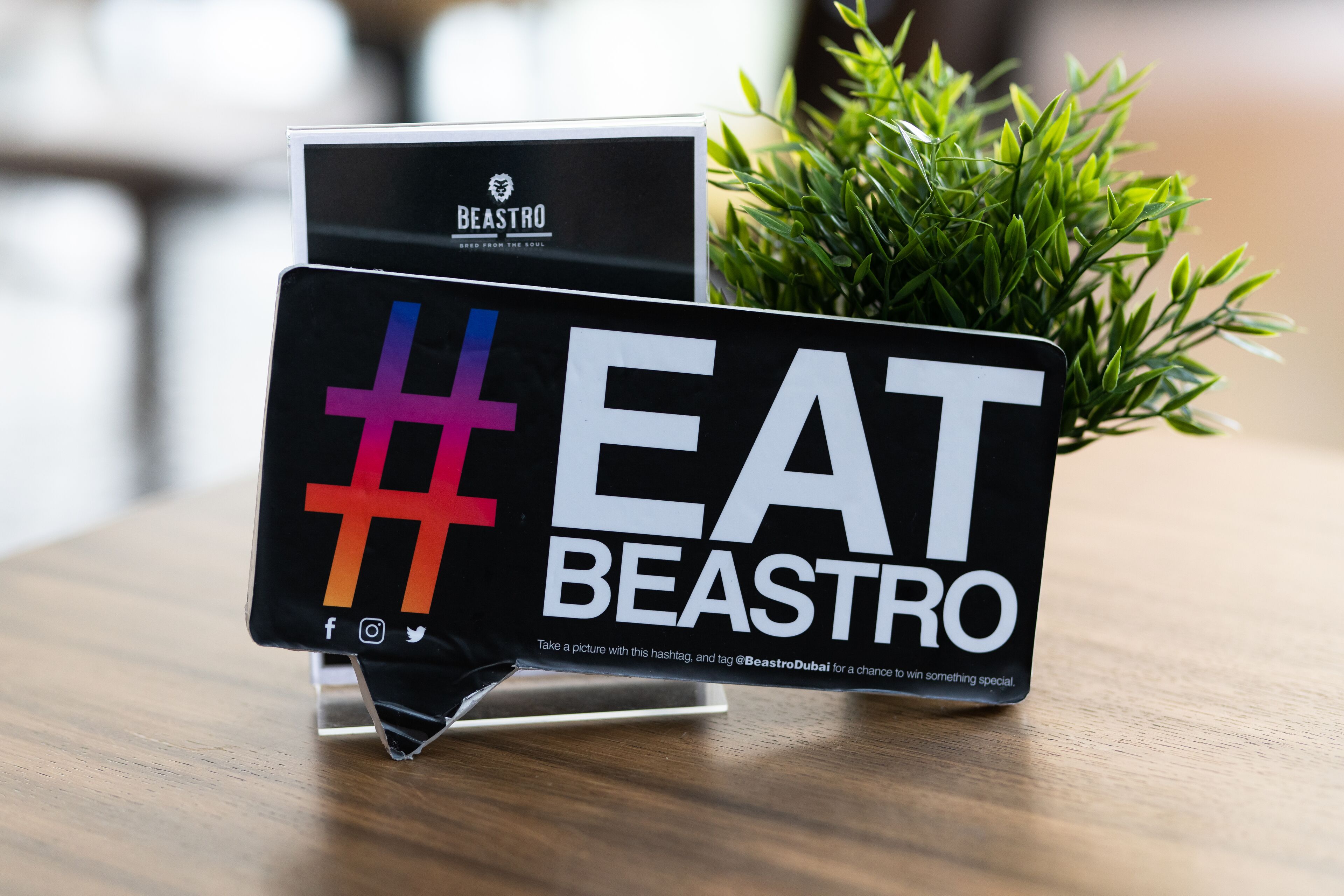 Reklameskilt for 'EAT BEASTRO' med hashtag og sociale medier-ikoner, placeret på et stativ med en lille potteplante i baggrunden.