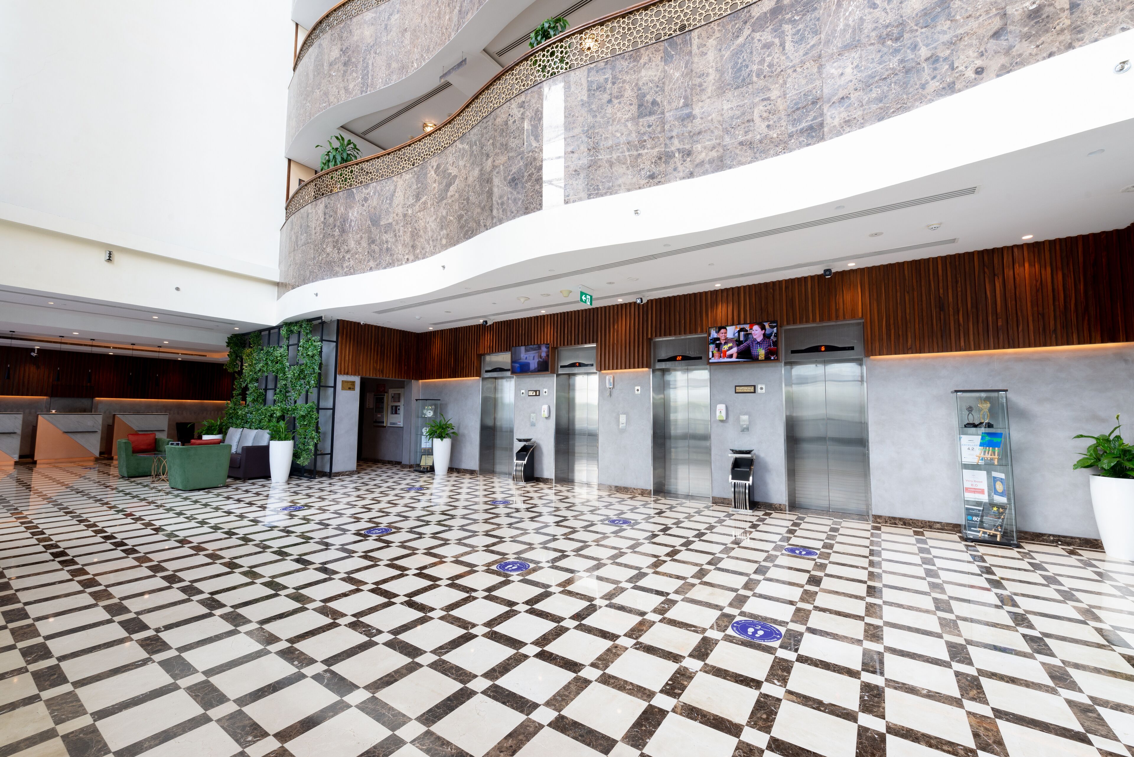 A spacious lobby with a checkered floor, multiple elevators, a reception area, and a high ceiling with a decorative wall.