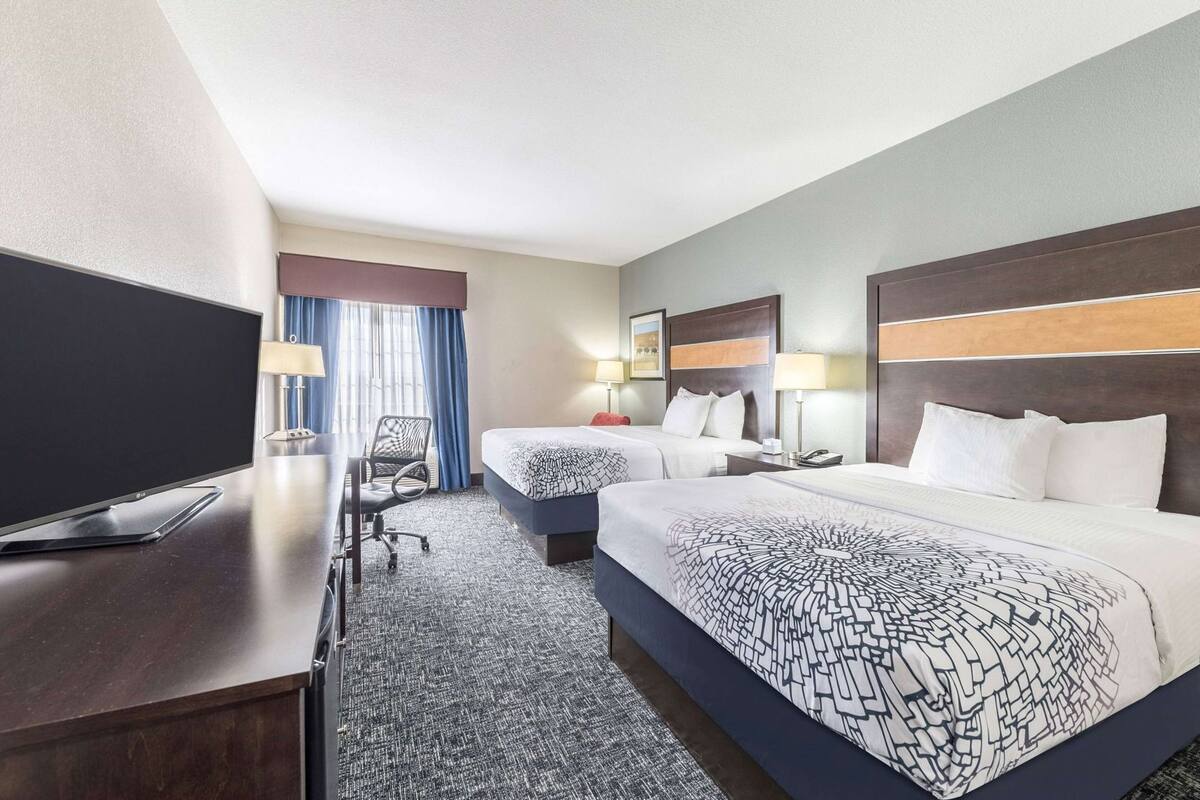 Ein Hotelzimmer mit zwei Betten, einem Schreibtisch, einem Flachbildfernseher, einem Sessel und einem Fenster mit Vorhängen.