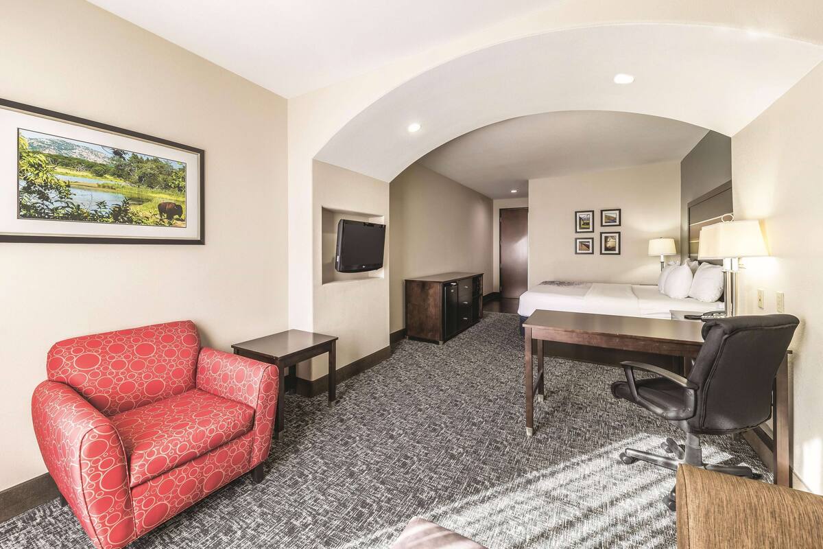 Ein Hotelzimmer mit einer roten, gemusterten Couch, einem Schreibtisch mit Stuhl, einem Bett, einem an der Wand montierten Fernseher und gerahmten Bildern.