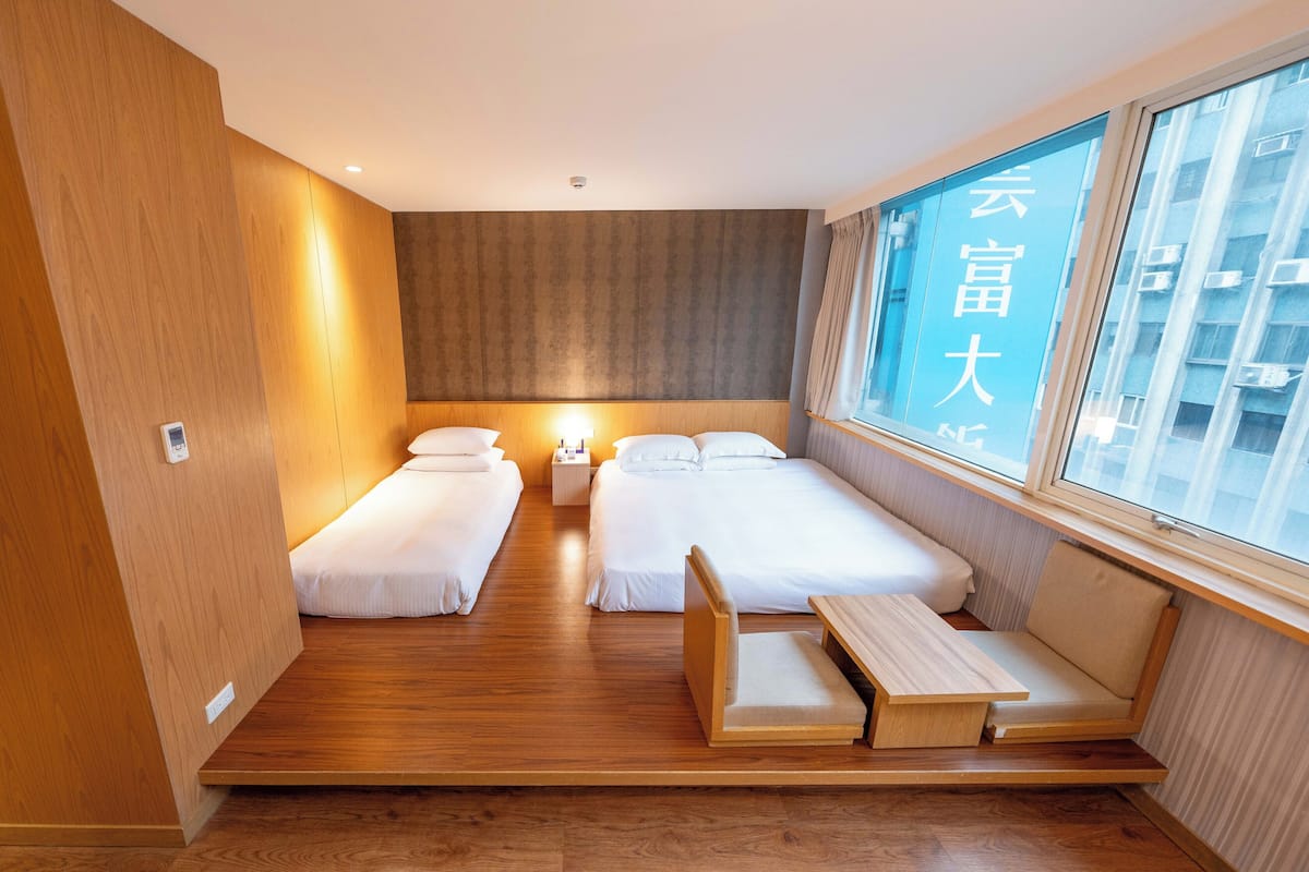A hotel room with two beds, a small table, and a bench.