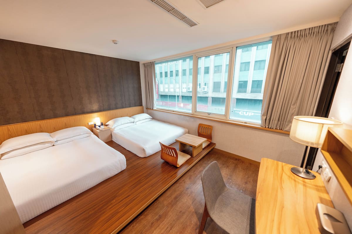 A hotel room with two beds, a desk, a chair, a small table, and a window with curtains.
