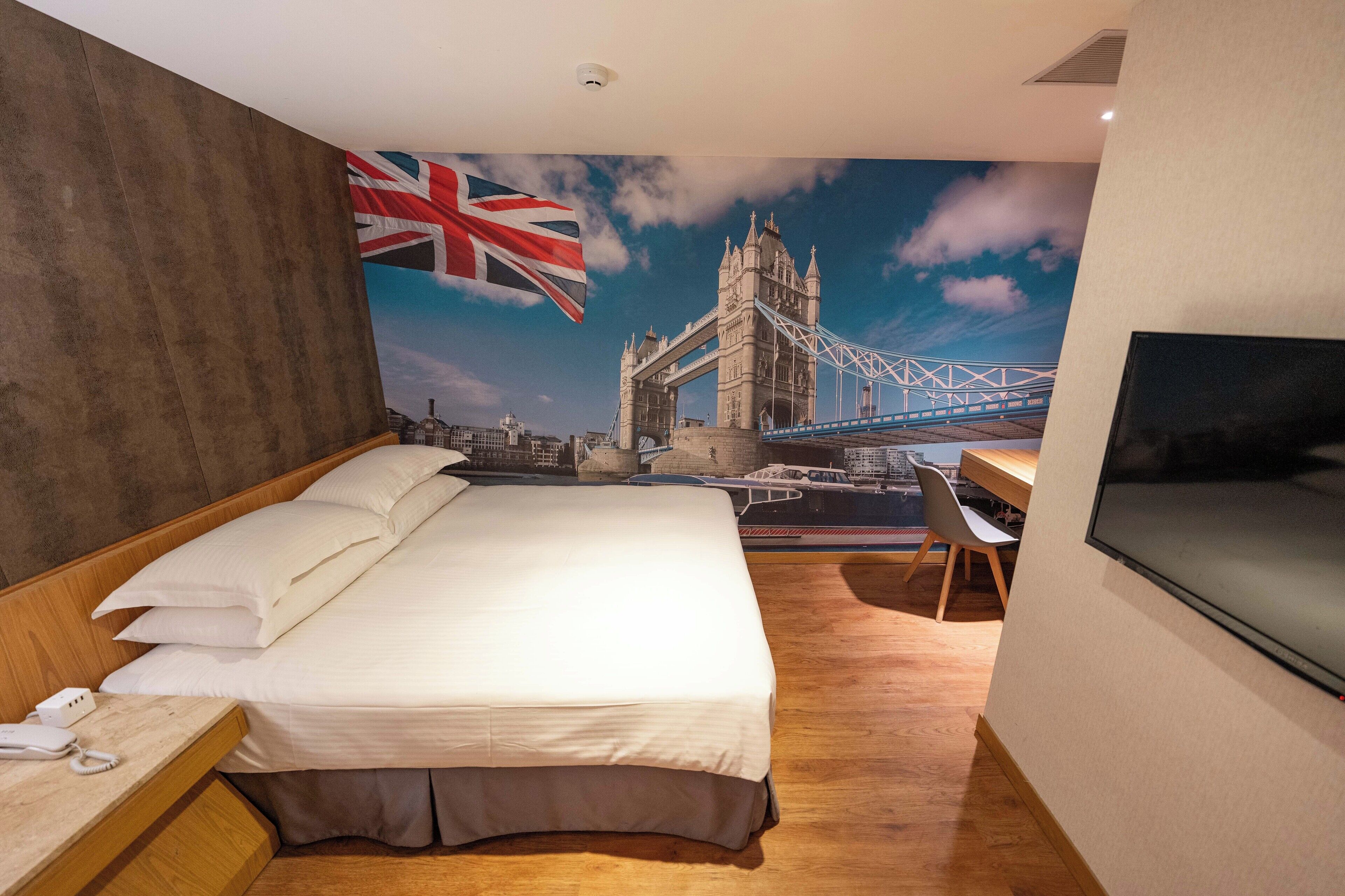 A hotel room with a large bed, a desk, a chair, and a TV. There is a mural of a bridge and a flag on the wall.