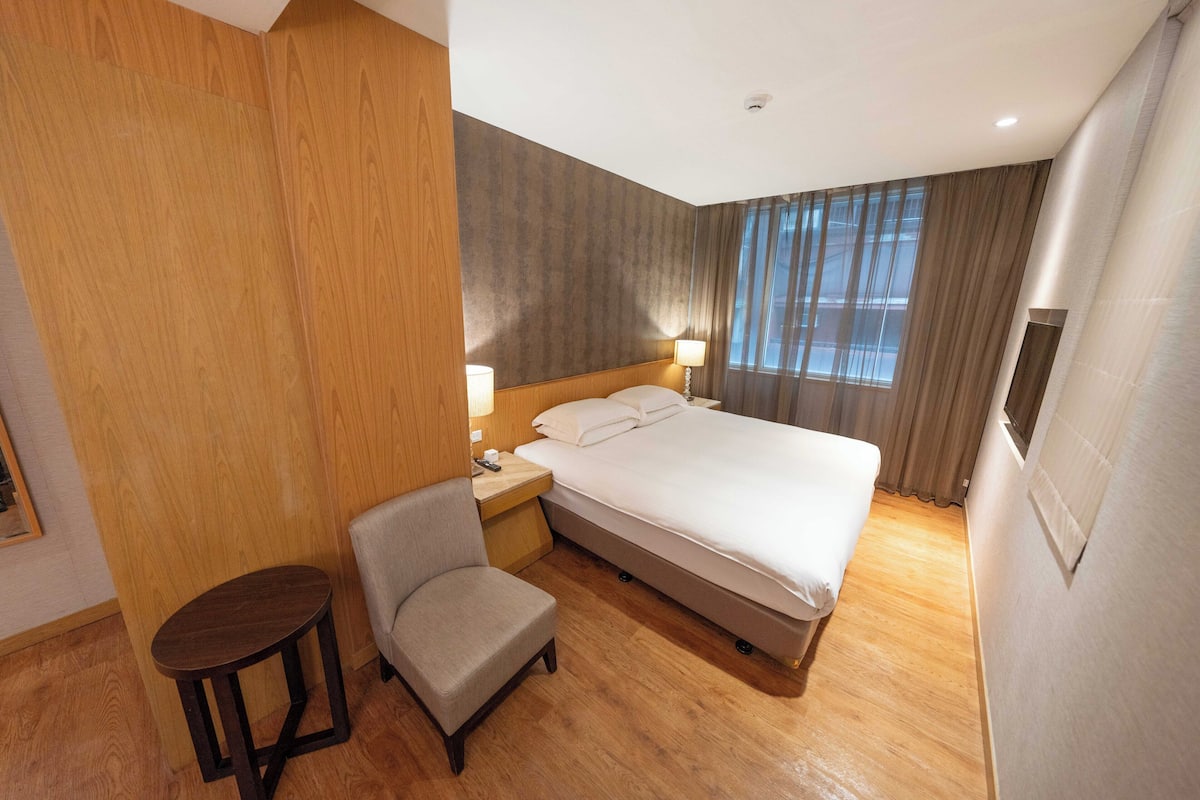 A hotel room with a bed, a chair, a small table, a nightstand, and a window with curtains.