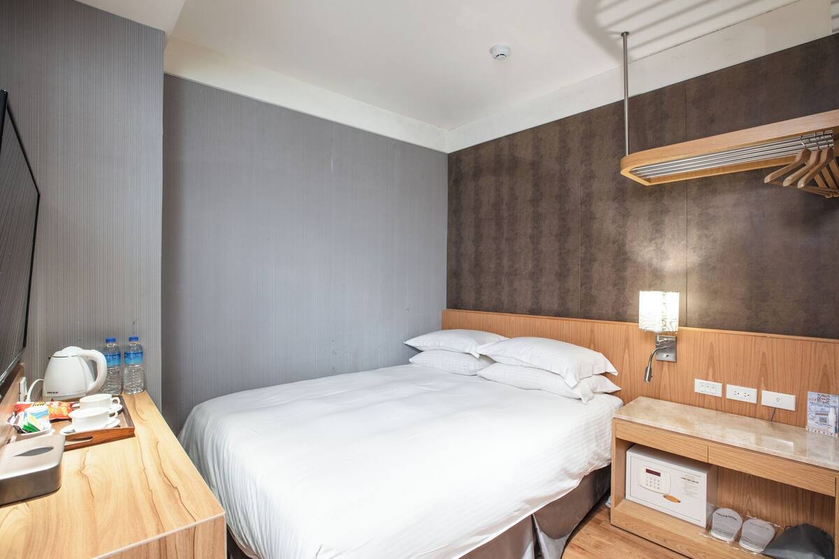 A hotel room with a large bed, a wooden desk with a kettle, cups, and a tray, and a wall-mounted heater.
