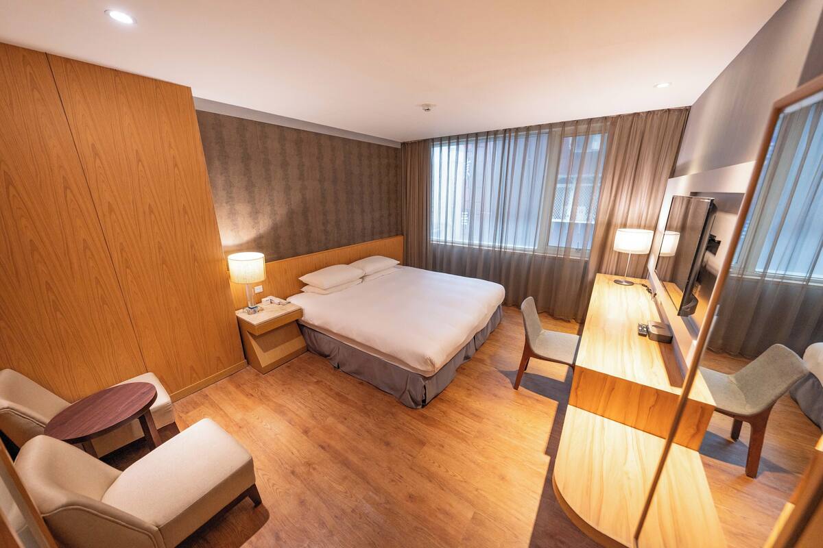 A hotel room with a large bed, a desk with a chair, a small table, and a mirror.