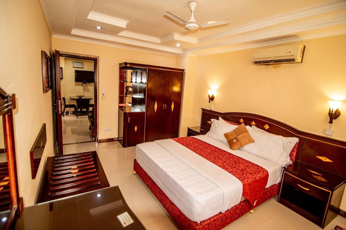 A hotel room with a bed, a desk, a wardrobe, a ceiling fan, and an air conditioning unit.