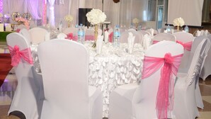 Un salón de banquetes con mesas redondas cubiertas con manteles blancos, sillas con fundas blancas y cintas rosas, y centros de mesa florales.