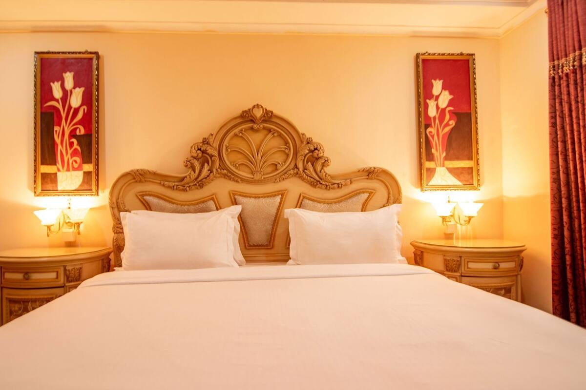 A hotel room with a large bed, ornate headboard, two bedside tables, and two wall-mounted lamps.
