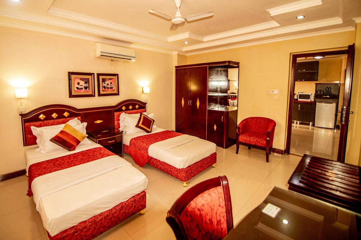 A hotel room with two beds, a desk, a chair, a wardrobe, and a kitchenette.
