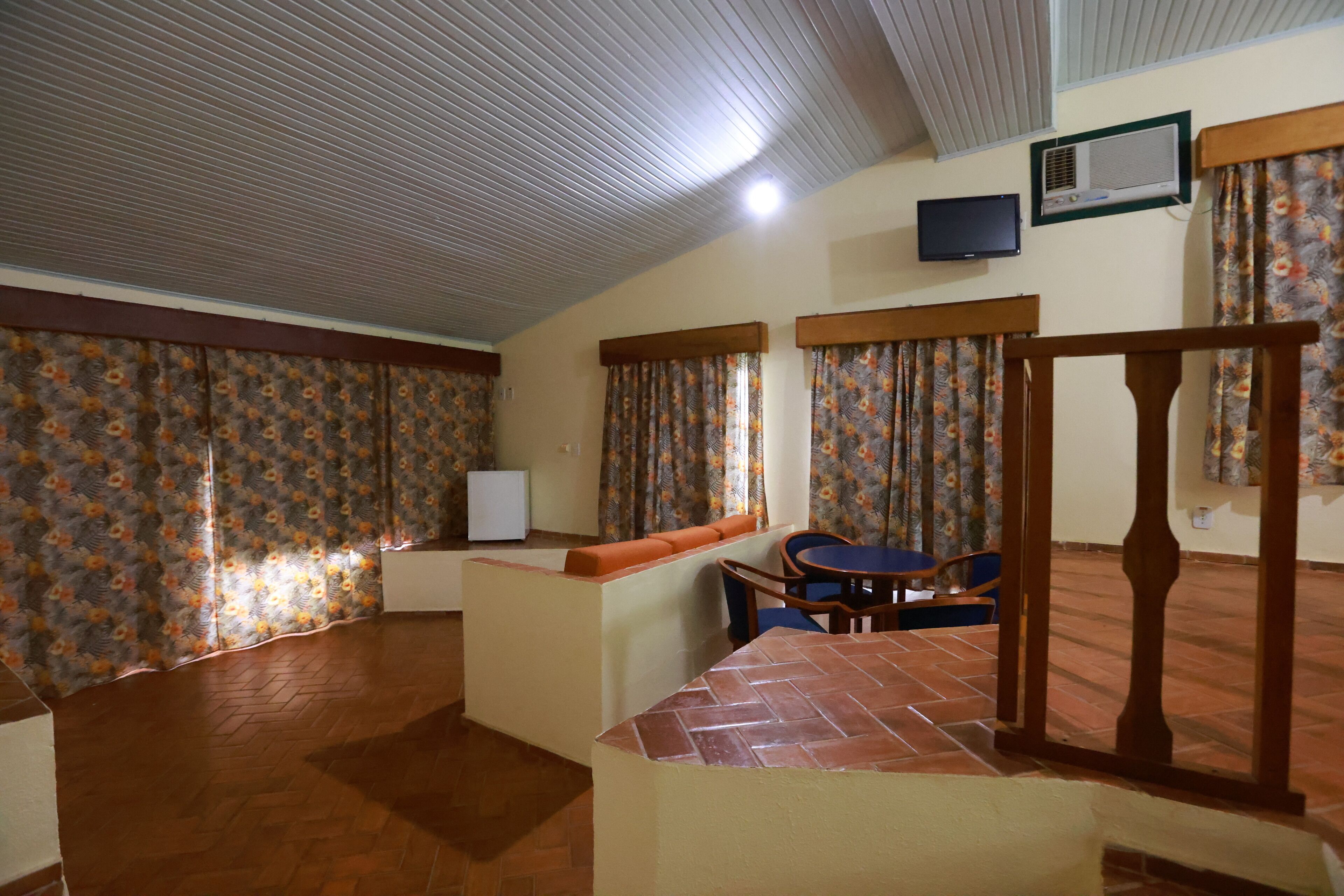 Um quarto com piso de azulejos, um sofá com almofada, uma mesa pequena, uma cadeira, uma televisão e uma janela com cortinas.