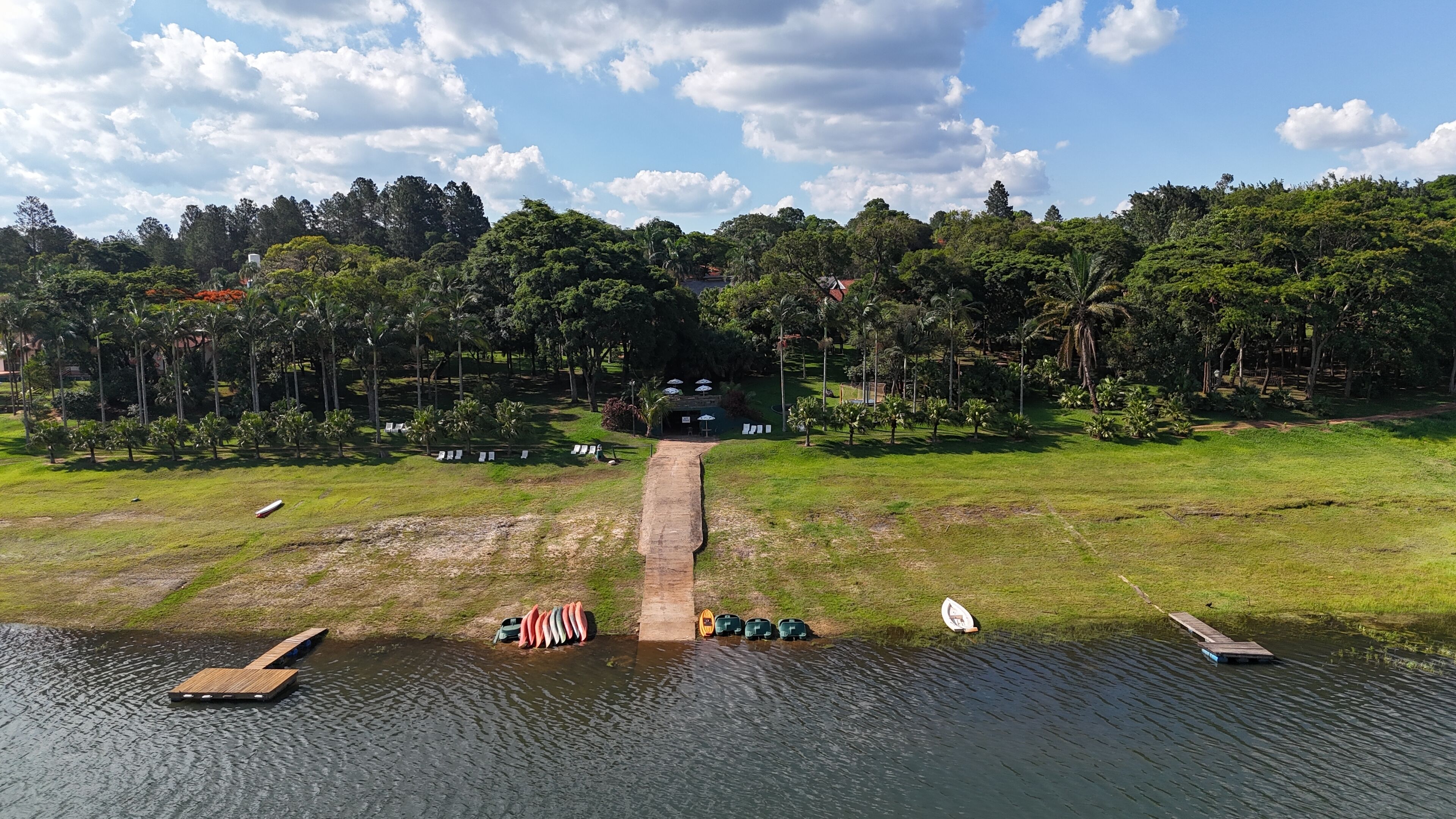 A lakeside view with a wooden dock, a row of canoes, and a pathway leading to a building surrounded by trees.