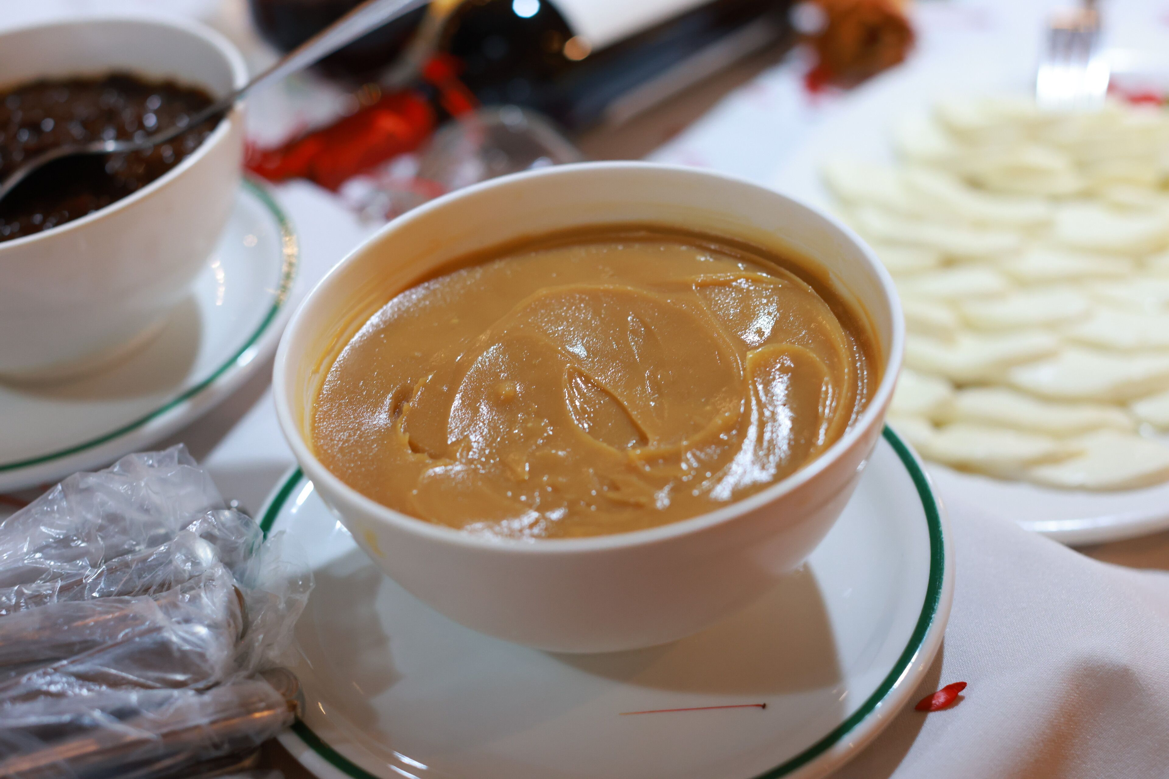 A bowl of soup on a white plate with a green rim, accompanied by a bowl of chili and a plate of sliced potatoes.