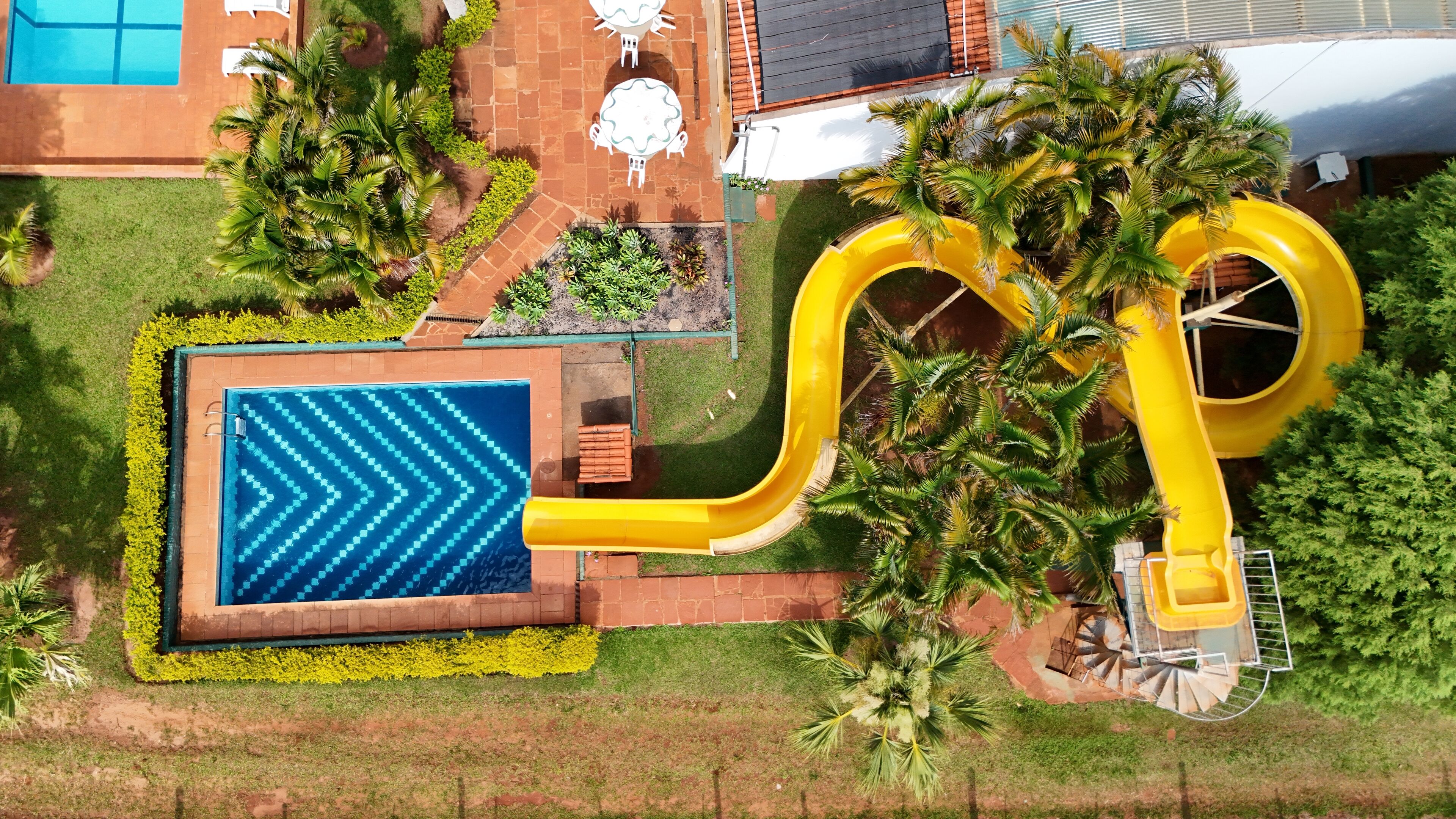 Vista aérea de uma área de piscina com um escorregador amarelo, uma piscina retangular com padrão em chevron e vegetação ao redor.