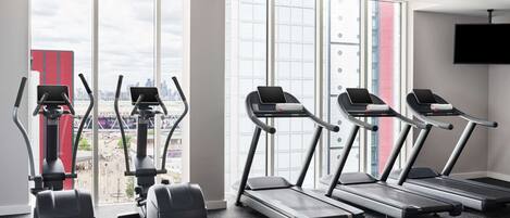 A gym with treadmills, elliptical machines, and a stationary bike, large windows with a city view, and a flat-screen TV mounted on the wall.