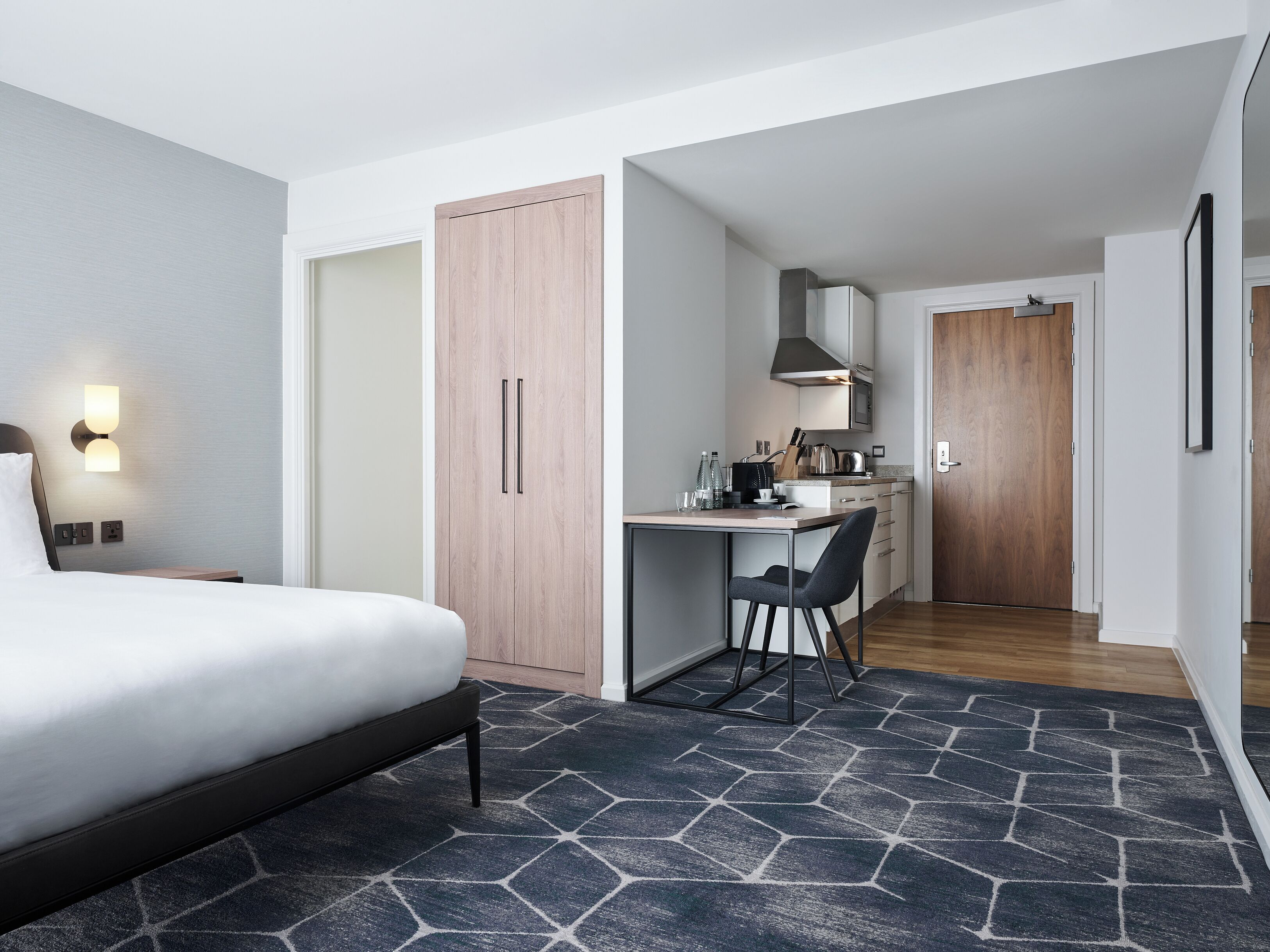 A hotel room with a bed, a desk, a chair, a kitchenette, and a wardrobe.