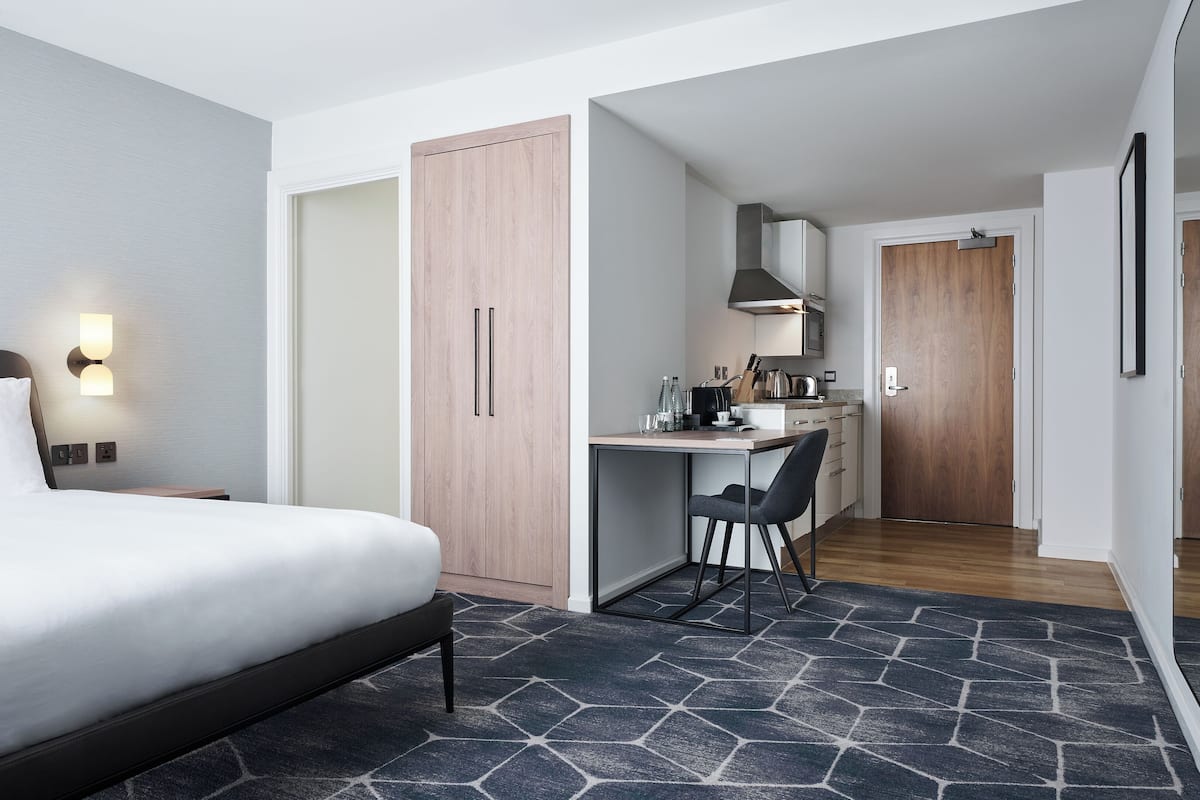 A hotel room with a bed, a desk, a chair, a kitchenette, and a wardrobe.