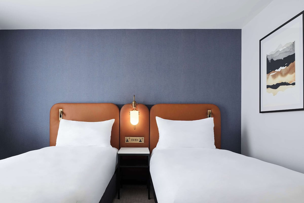 A hotel room with two single beds, a nightstand, a wall-mounted lamp, and a framed picture on the wall.